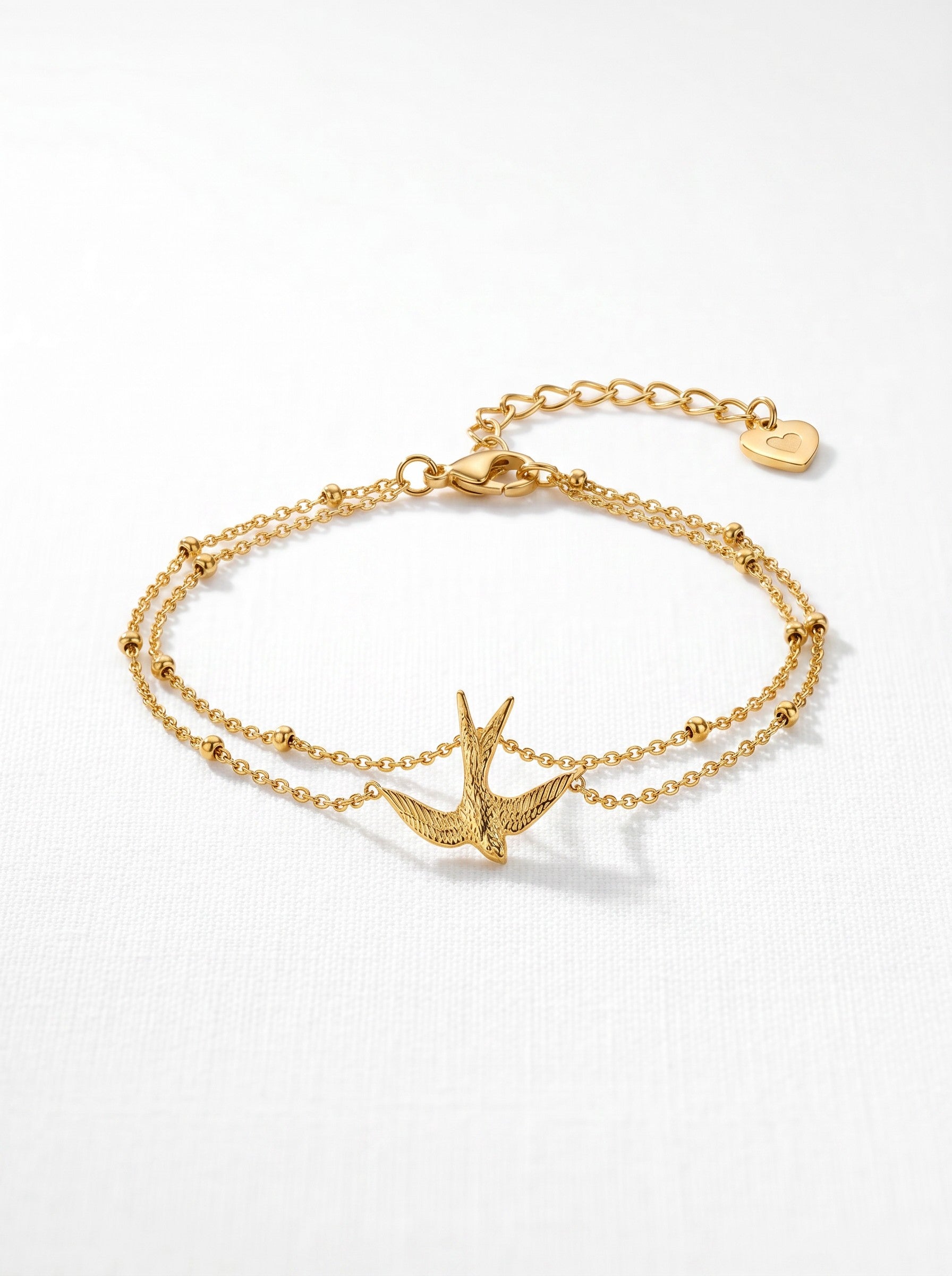 Color's Wholesale gold double-chain bracelet featuring a textured swallow bird charm and a small heart pendant.