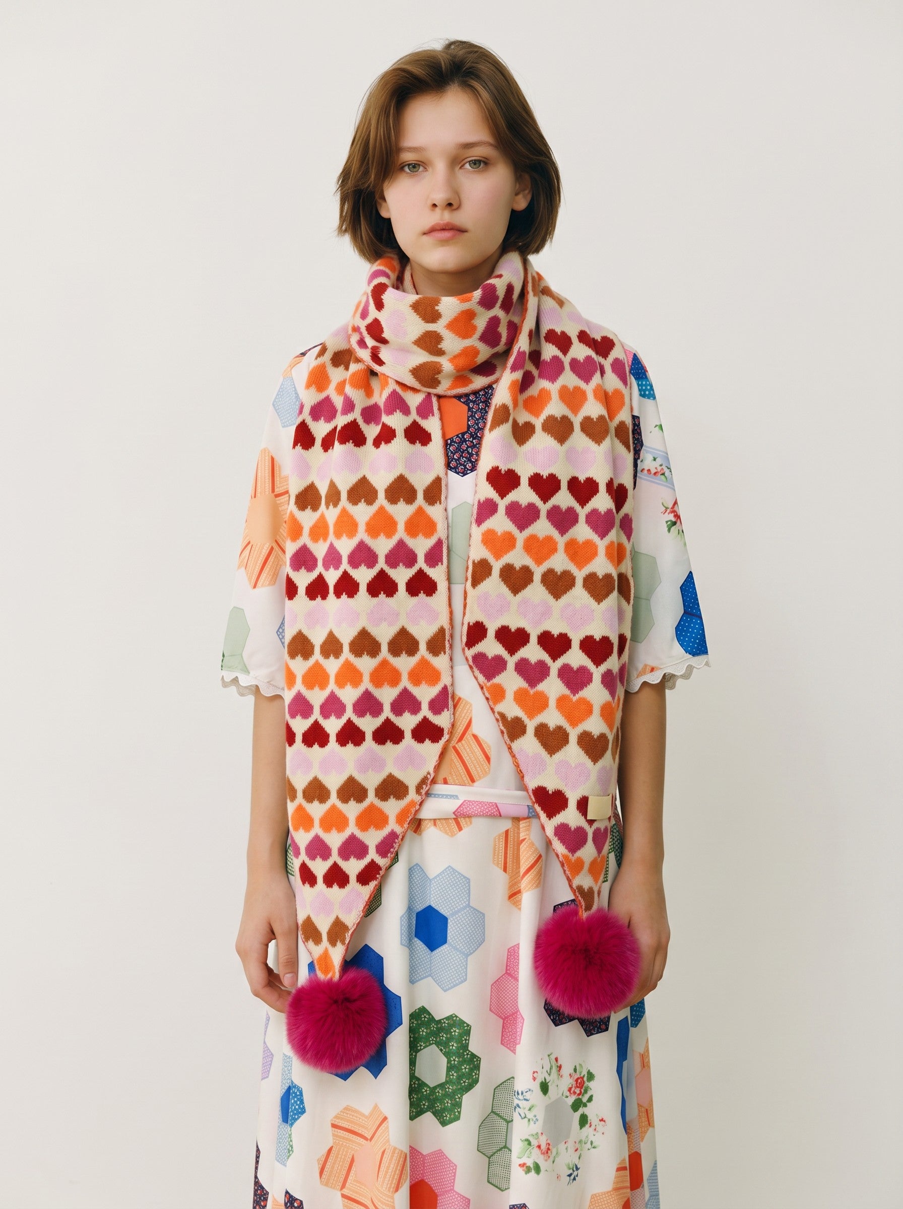 Color's Wholesale knit scarf featuring a multi-colored heart pattern with pink faux fur pom-poms on the ends.