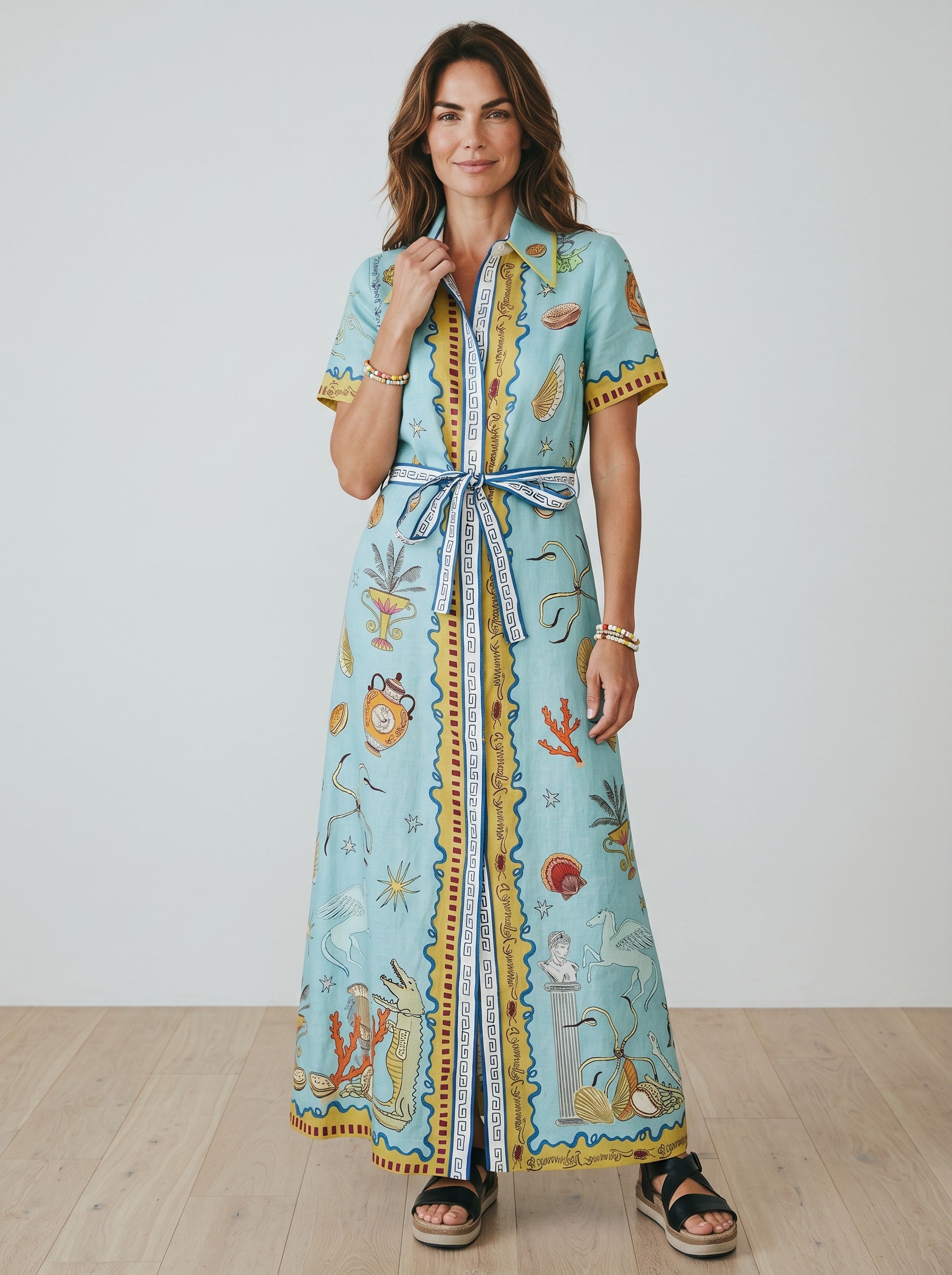 Color's Wholesale light blue linen-blend maxi dress with a bold print of mythological figures, shells, and coral. Features short sleeves, a belted waist, and a collared neckline.