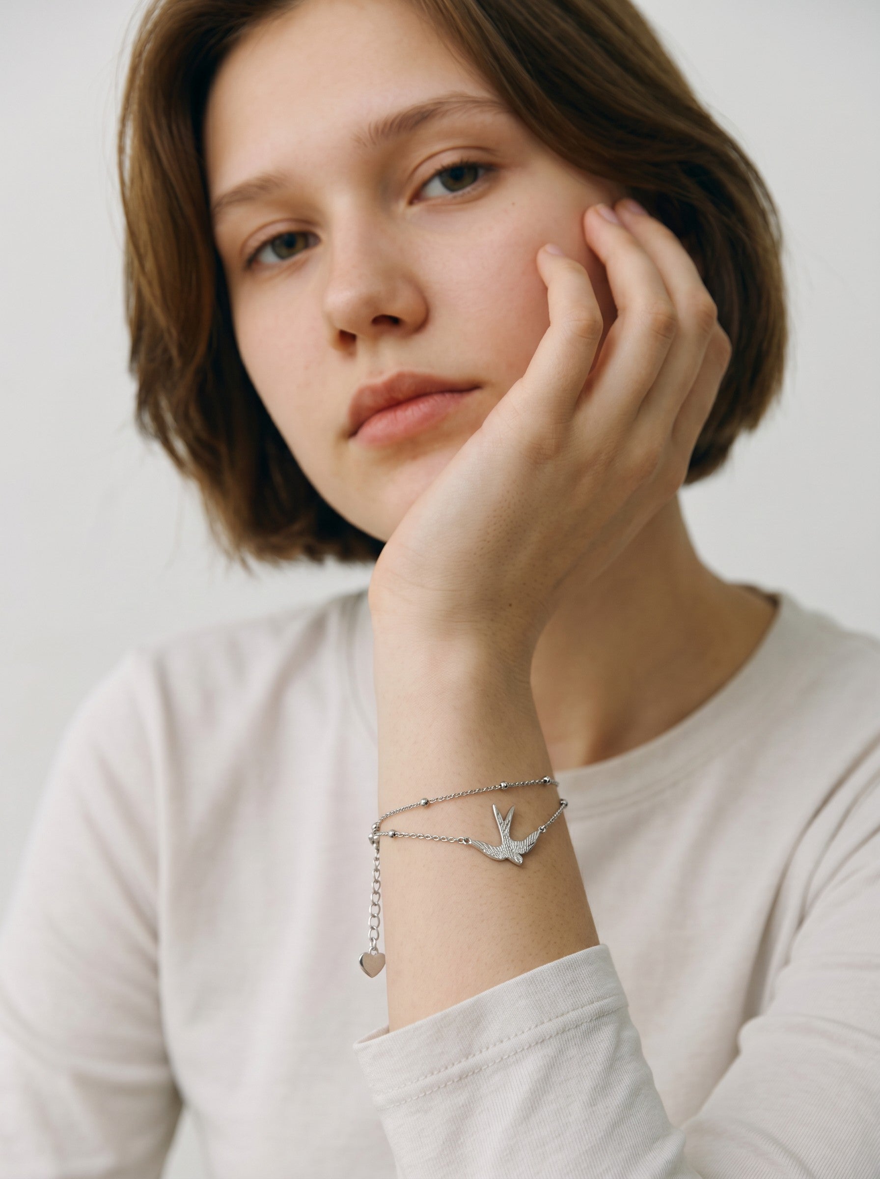 Color's Wholesale silver bird charm bracelet on a woman's wrist, featuring a delicate double chain and heart charm.