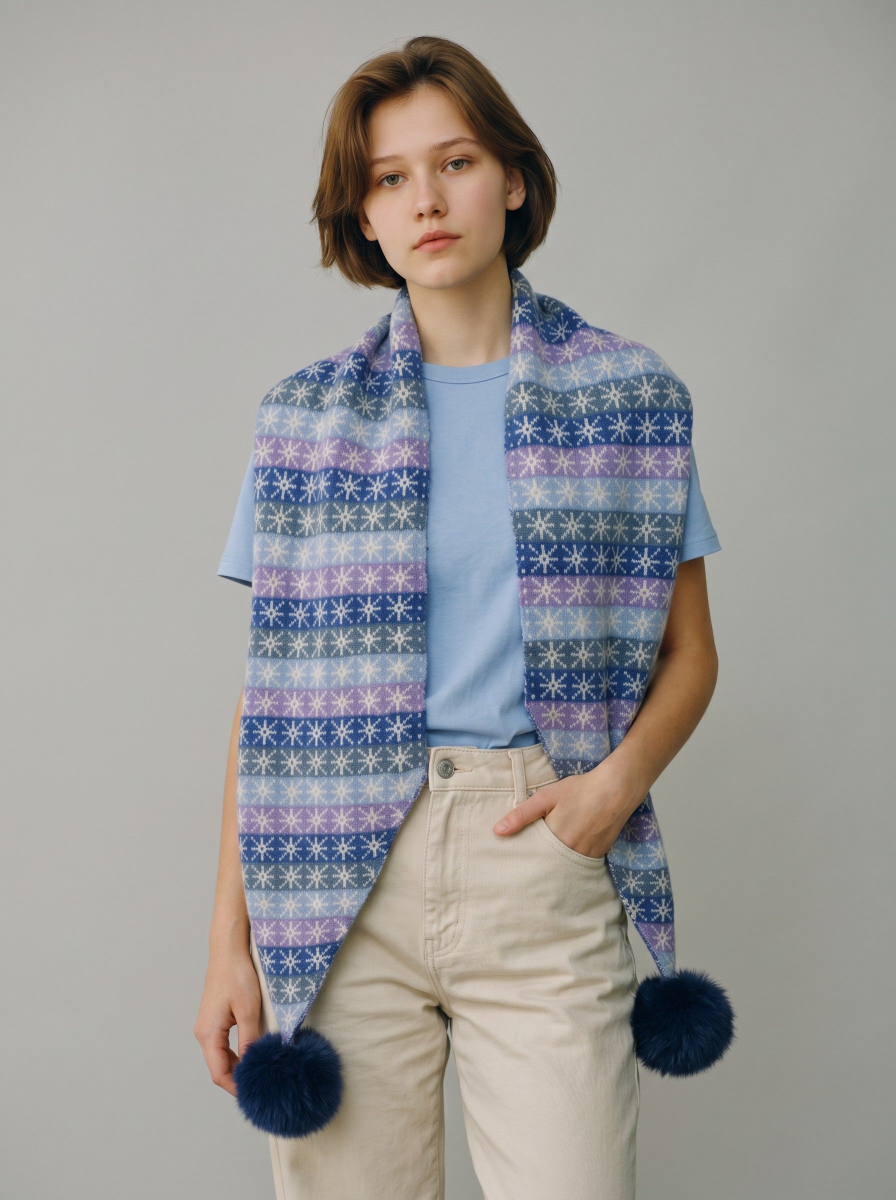 Color's Wholesale blue and purple snowflake patterned knit scarf with fluffy pom-poms worn by a woman.